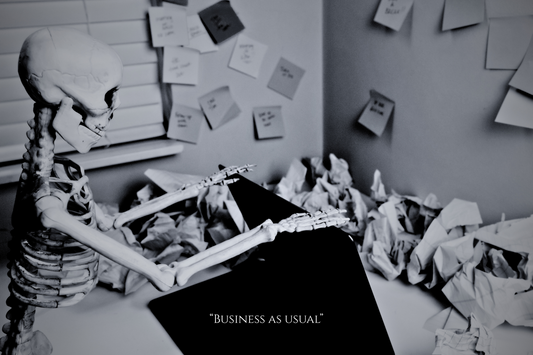 Black-and-white image of a plastic skeleton sitting at a cluttered desk with a laptop, sticky notes, paper stacks, and a pen in hand — a humorous visual metaphor for late-night editing fatigue.
